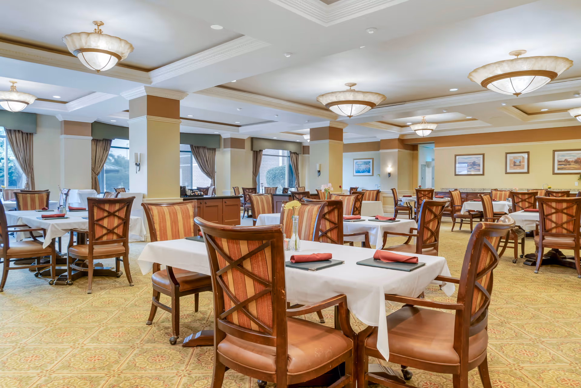 A spacious dining room with multiple tables covered in white tablecloths, each set with red napkins and menus. The room features wooden chairs with striped upholstery, large windows with curtains, beige walls with framed artwork, and elegant ceiling light fixtures.