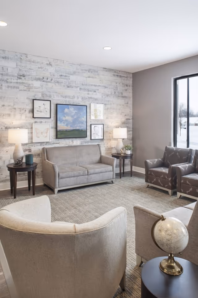 Comfortable living room with a gray sofa and armchairs arranged around side tables, wall art, lamps, and a window showing snow outside.