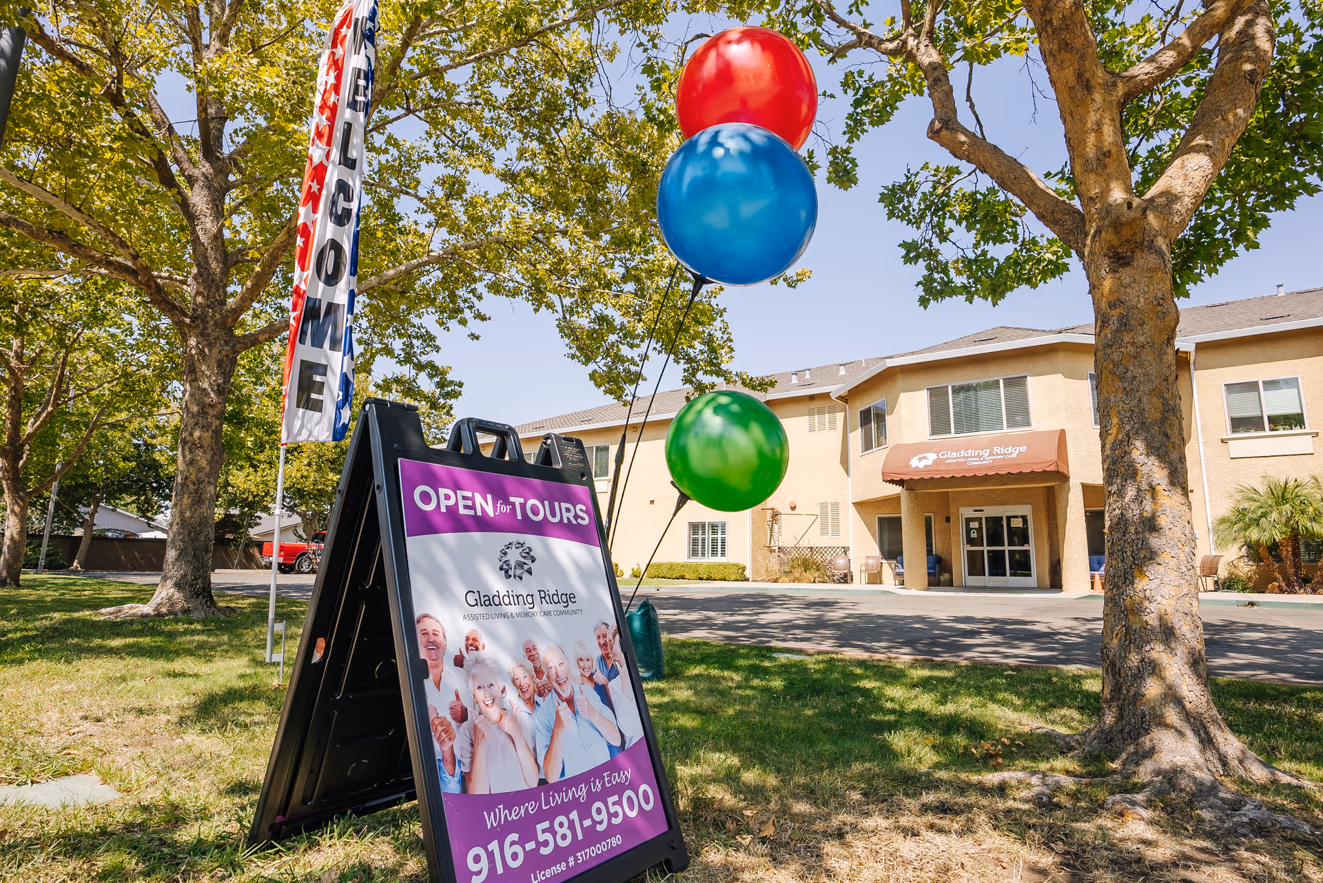 Outdoor view of Gladding Ridge Assisted Living and Memory Care facility in Lincoln, California, showing the building entrance with an awning, a signboard advertising open tours with smiling seniors, colorful balloons, and a welcome flag among trees on a sunny day.