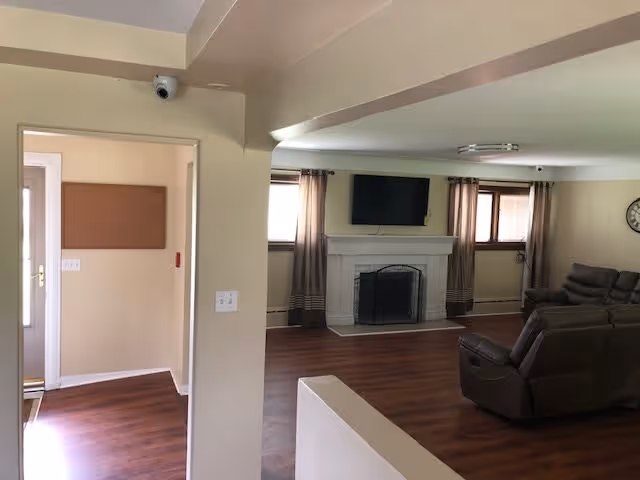 Interior view of a senior living facility common area with wooden flooring, a white fireplace with a mounted flat-screen TV above it, two windows with curtains on either side of the fireplace, a wall clock, and brown cushioned recliner chairs arranged in the room.