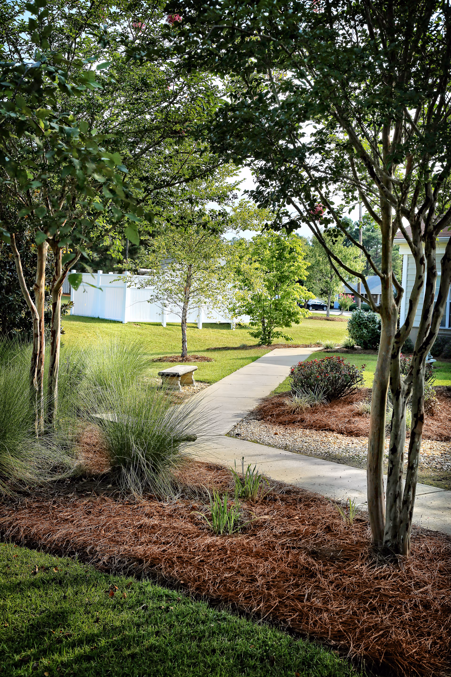 Curved concrete walkway winds through landscaped grounds with trees, shrubs, a bench, and a white fence in the background.