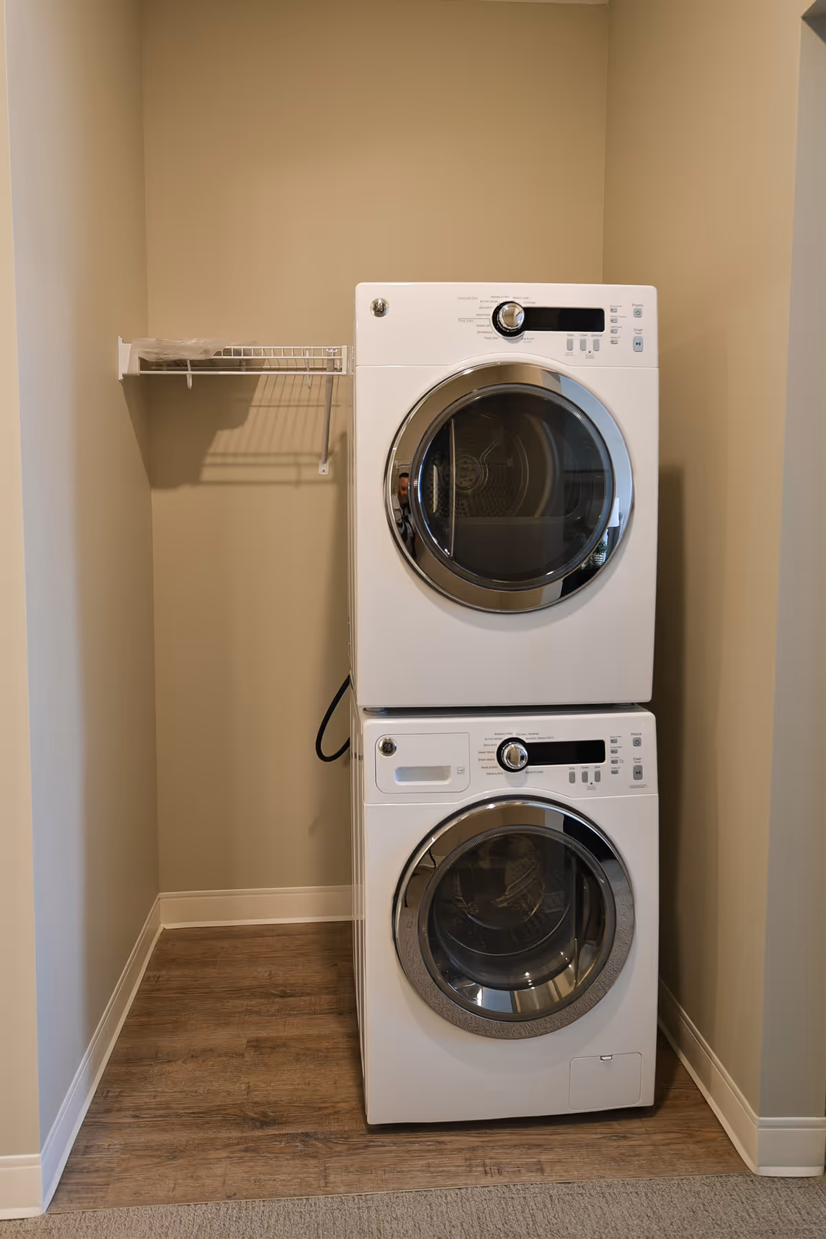 A small laundry area with a stacked white front-loading washer and dryer set against beige walls and wood flooring. There is a white wire shelf mounted on the left wall above the machines.
