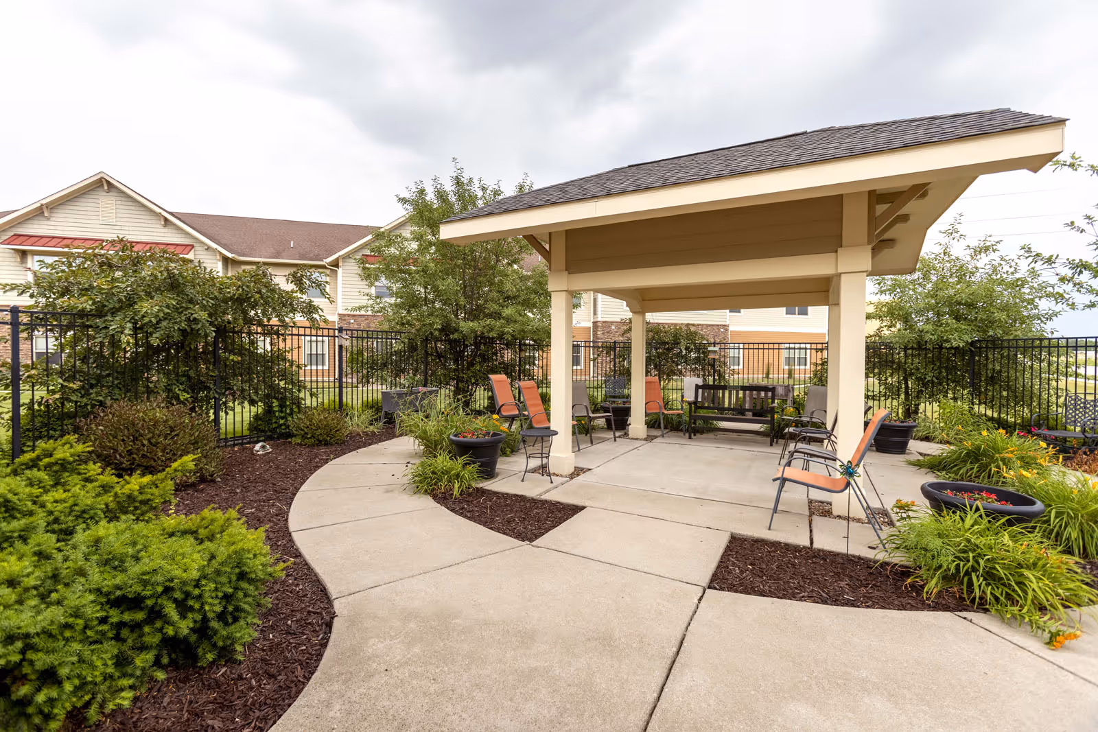 Outdoor seating area with a covered pavilion featuring several chairs and a bench, surrounded by landscaped plants and bushes. A black metal fence encloses the area, and a multi-story residential building is visible in the background under a cloudy sky.