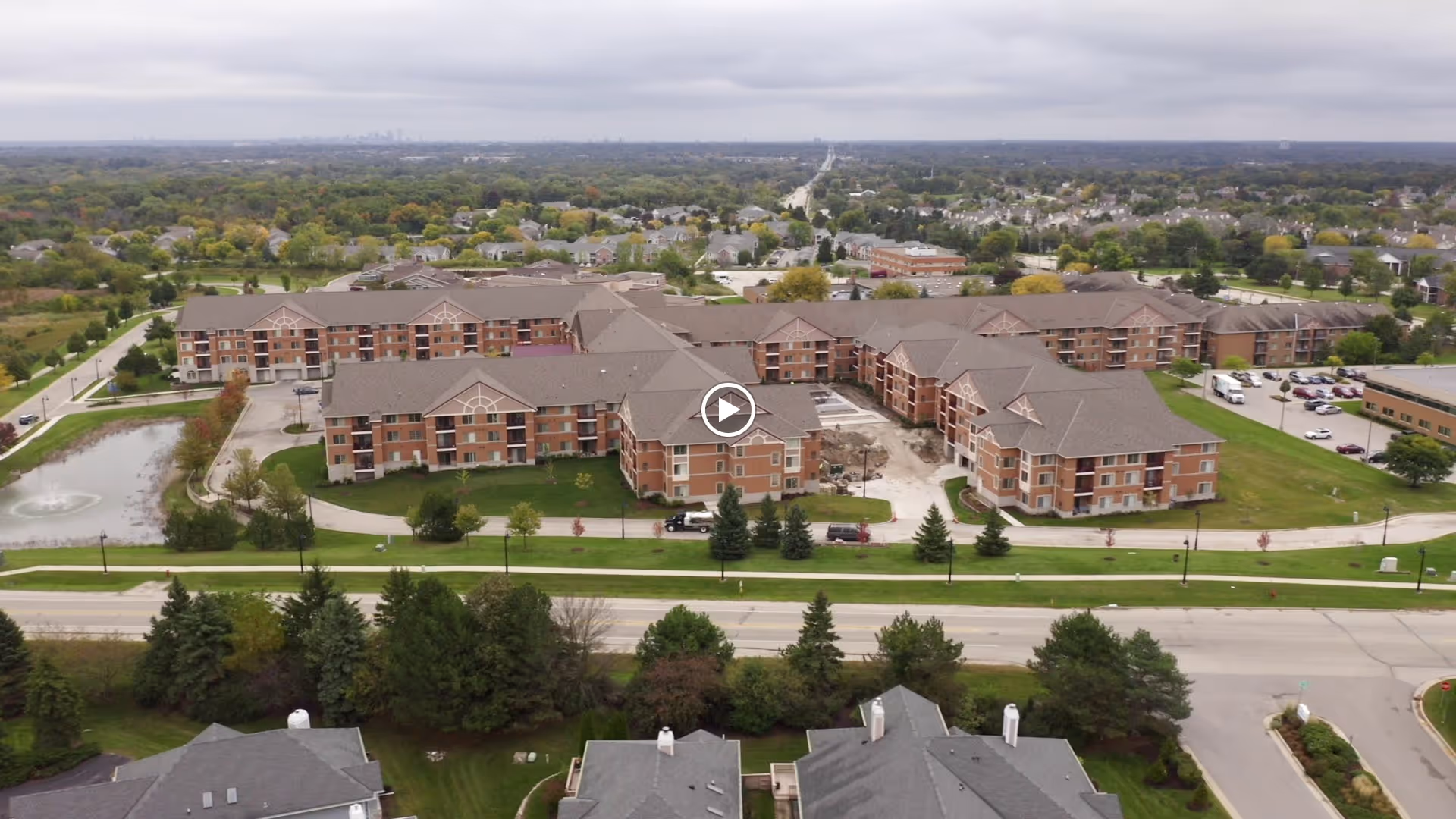 Aerial view of ProHealth Regency Senior Communities New Berlin, showing multiple connected brick buildings with brown roofs surrounded by green lawns, trees, and a small pond with a fountain. Roads and residential neighborhoods are visible in the background under a cloudy sky.