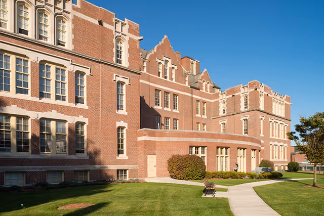 Red brick multi-story institutional building with decorative windows, a curved entrance area, a walkway and manicured lawn under a clear blue sky.