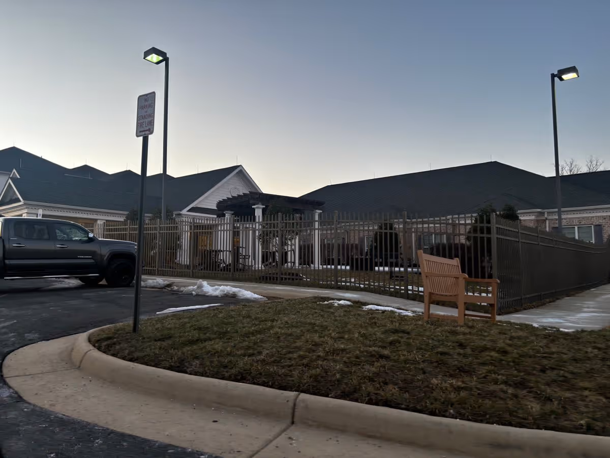 Exterior front of a senior living facility showing a fenced courtyard, bench, lamp posts, and a parked truck at dusk.