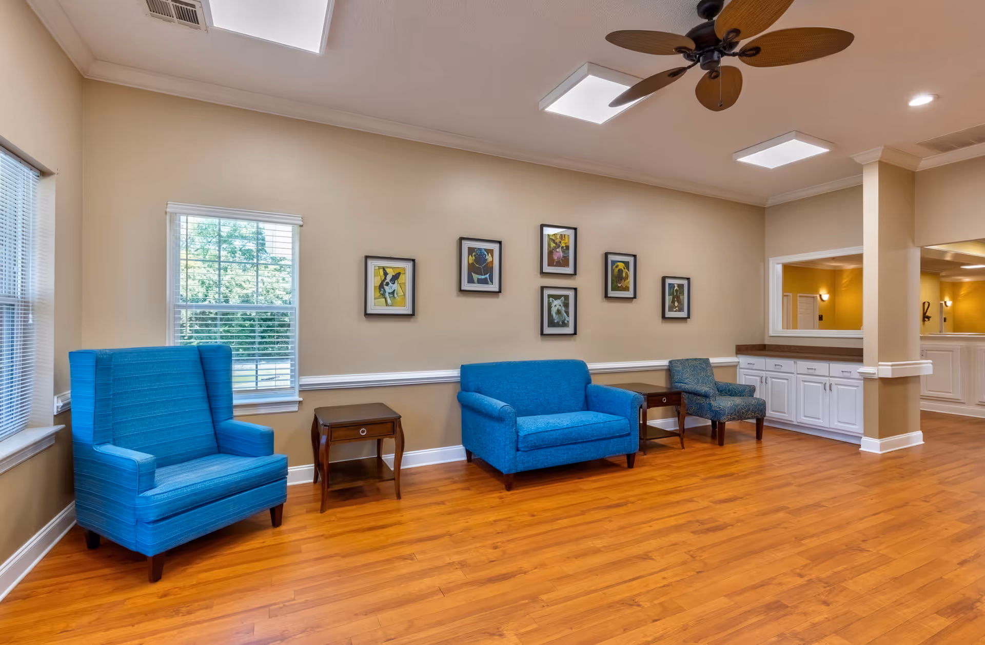 A bright and spacious living room area with wooden flooring, beige walls, and a ceiling fan. The room features a blue armchair, a blue loveseat, and a patterned blue chair, each separated by small wooden side tables. There are six framed pictures of dogs on the wall above the loveseat. Two windows with white blinds allow natural light into the room. A built-in white cabinet with a countertop is visible in the background.