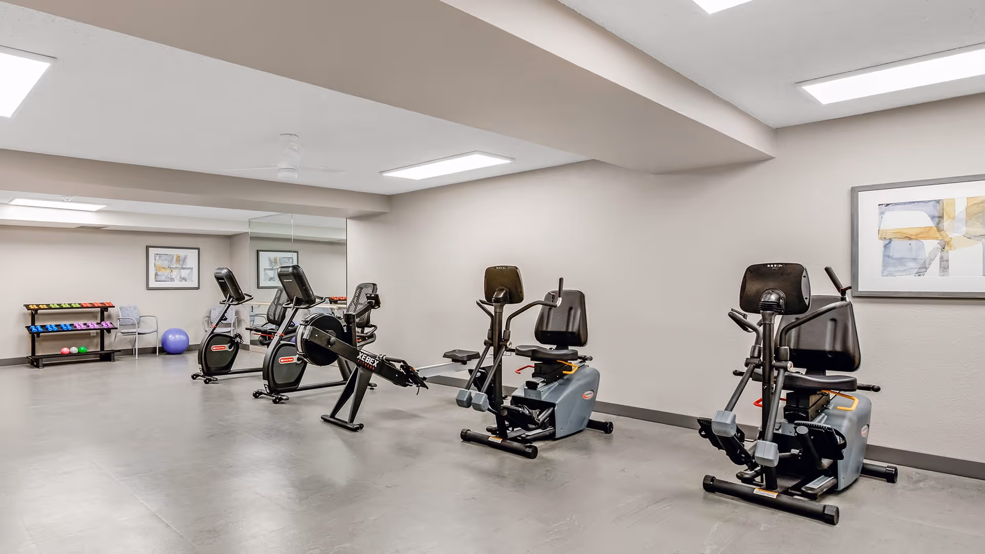 A fitness room in a senior living facility with exercise equipment including two recumbent bikes, two upright stationary bikes, and a rowing machine. There is a rack with colorful dumbbells and exercise balls in the background, along with a chair and framed artwork on the walls. The room has a light gray floor and neutral-colored walls with a large mirror on one side.