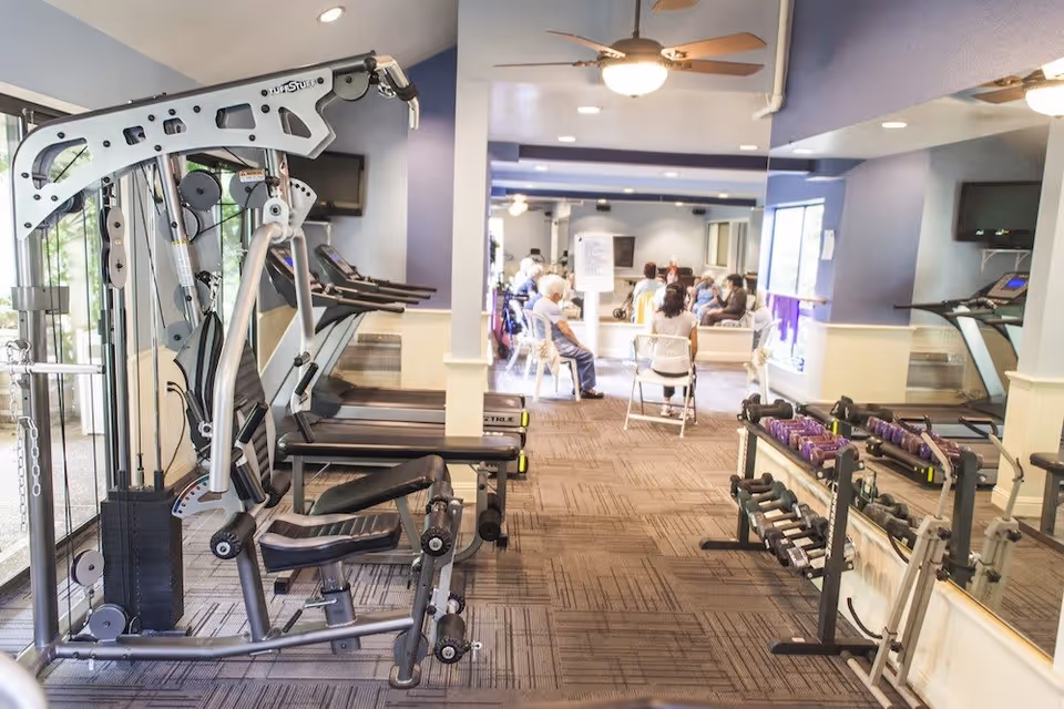 A fitness room with exercise equipment including a multi-gym machine, treadmills, and racks of dumbbells. In the background, several elderly people are seated in a group setting, possibly participating in a class or social activity. The room has large windows, ceiling fans, and mirrors along one wall.