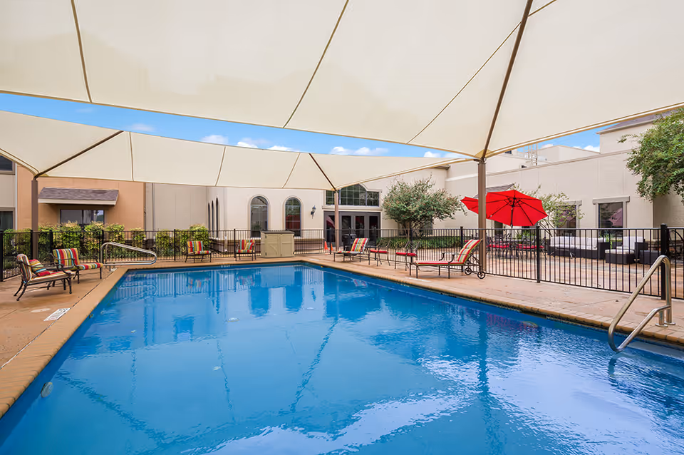 Outdoor swimming pool area at Villa de San Antonio with a large white canopy overhead, several lounge chairs with colorful cushions around the pool, a red umbrella, and a seating area with outdoor furniture. The pool is surrounded by a black metal fence and the building is visible in the background under a blue sky with some clouds.