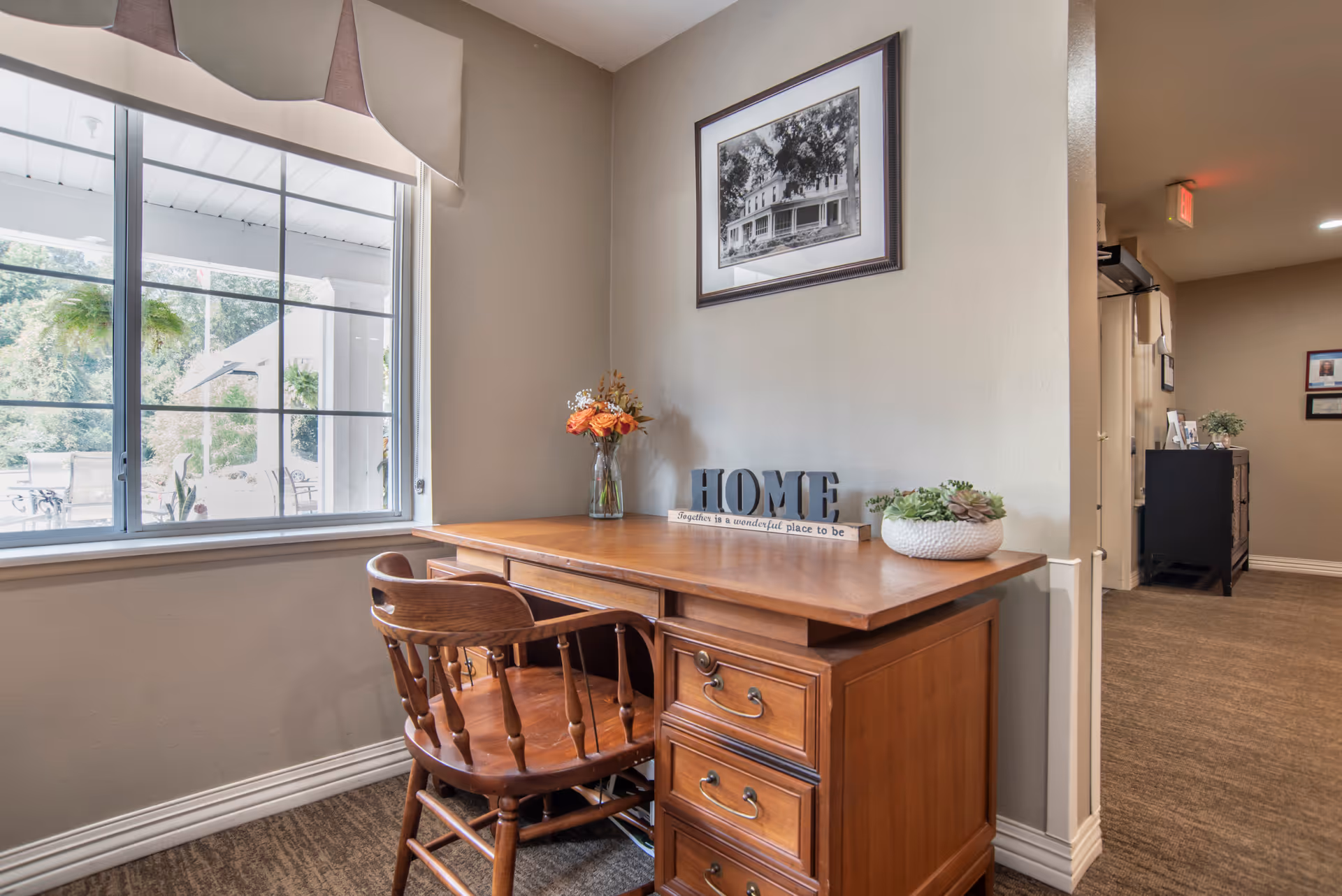 A cozy corner with a wooden desk and chair next to a large window showing an outdoor patio area. On the desk are a vase with orange flowers, a decorative sign that reads 'HOME', and a small potted succulent. A black and white framed photograph hangs on the beige wall above the desk. The scene is warmly lit with a carpeted floor and a hallway visible in the background.