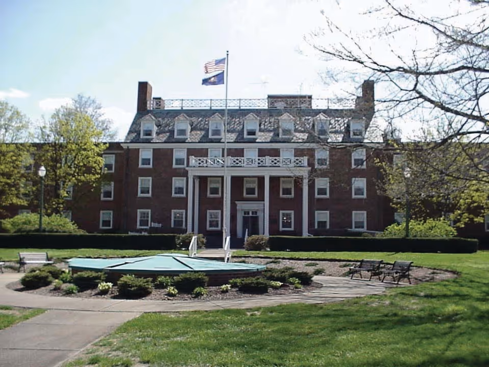 Front exterior view of a large brick building with multiple windows and dormers on the roof. There are two flagpoles with flags in front of the building, surrounded by a circular garden area with shrubs and benches. Trees and a grassy lawn are visible around the building under a clear sky.