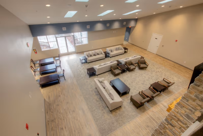 Spacious living room area with multiple beige sofas, armchairs, and coffee tables arranged on a large patterned rug. There are also several wooden tables with chairs near large windows and glass doors letting in natural light. The room has light wood flooring, beige walls, and a stone fireplace on the right side.