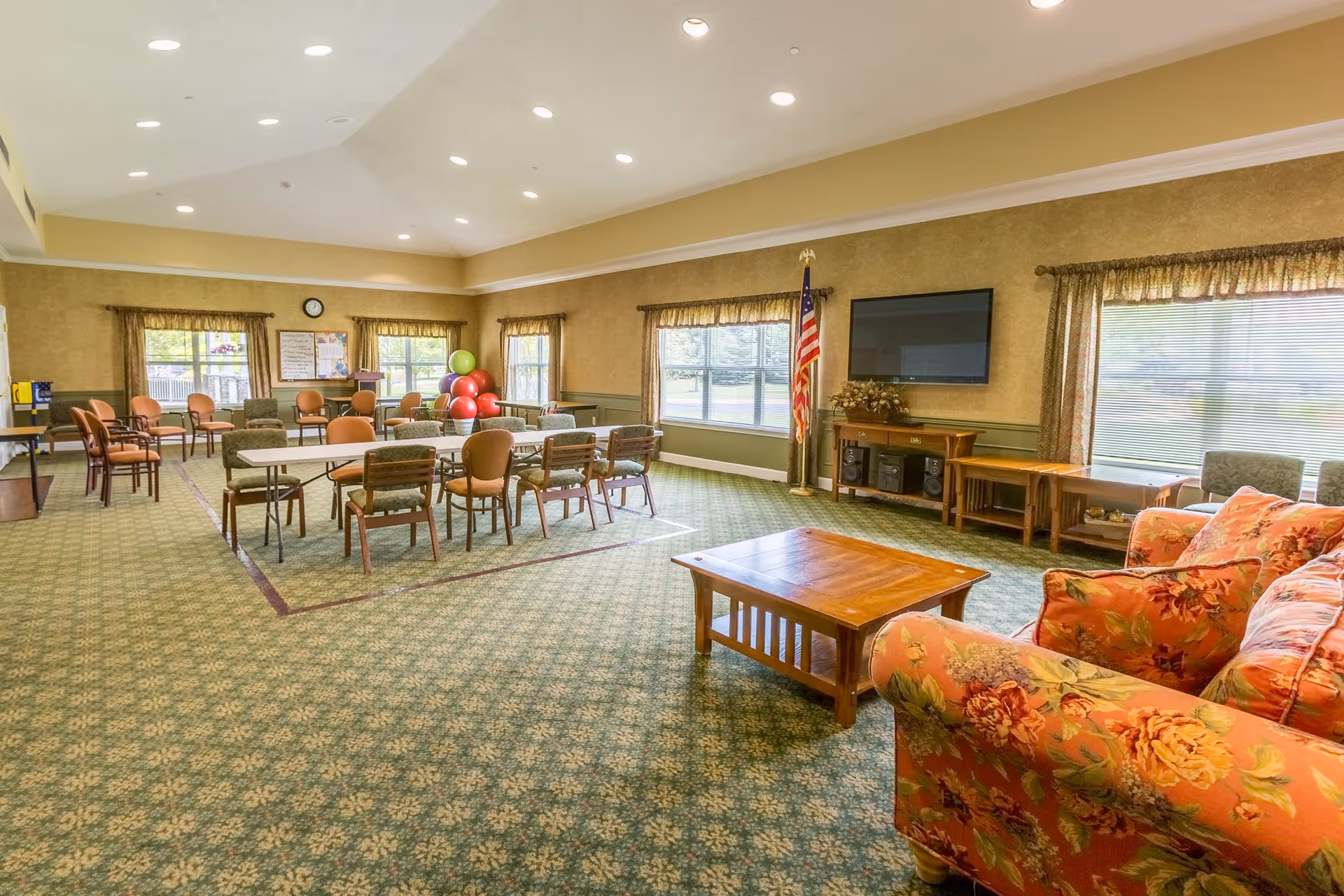 A spacious common room with patterned green carpet and beige walls. The room features several tables and chairs arranged for group activities or dining. There is a floral-patterned sofa and wooden coffee table in the foreground. Large windows with curtains allow natural light to fill the room. An American flag stands next to a flat-screen TV mounted on the wall above a wooden cabinet. Exercise balls and a bulletin board are visible in the background.