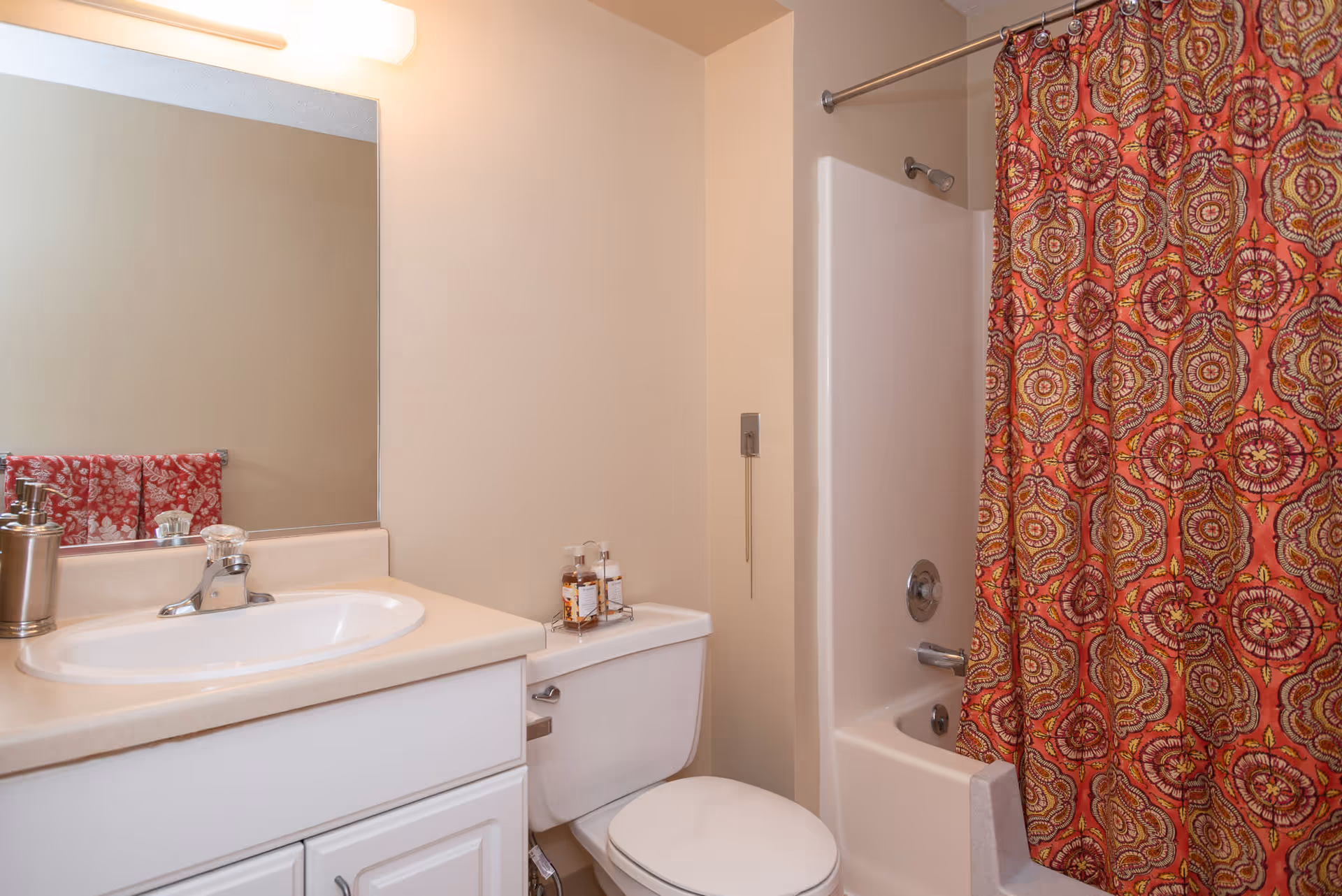 Bathroom with a white sink and countertop, a large mirror above the sink, a toilet with two bottles of soap or lotion on top, and a bathtub with a colorful red and orange patterned shower curtain.