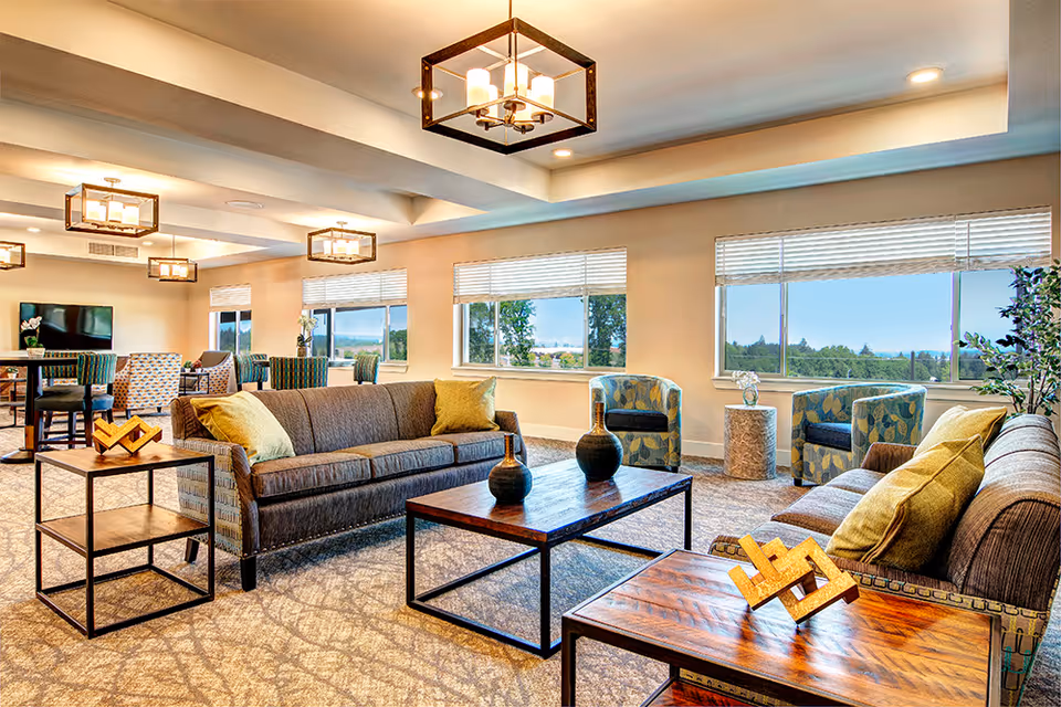 A bright and spacious senior living community lounge area with large windows showing a scenic outdoor view. The room features two brown sofas with yellow pillows, two patterned armchairs, a wooden coffee table with decorative vases, and side tables with geometric decor. Modern square pendant lights hang from the ceiling, and there are additional seating areas with chairs and tables in the background.
