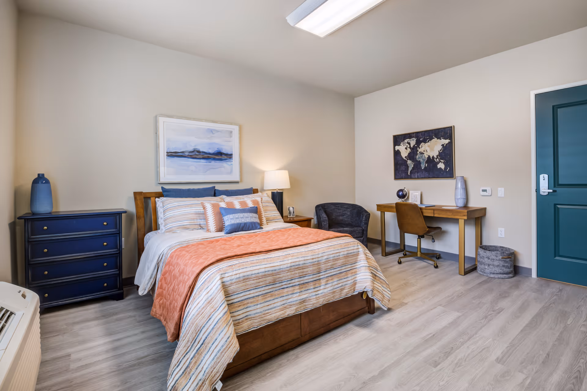 A neatly arranged bedroom in a senior living facility featuring a bed with striped bedding and multiple pillows, a wooden nightstand with a lamp, a blue chest of drawers with a decorative vase, a small desk with a chair, a globe, and a framed world map on the wall. The room has light-colored walls and flooring, and a teal door is visible on the right side.