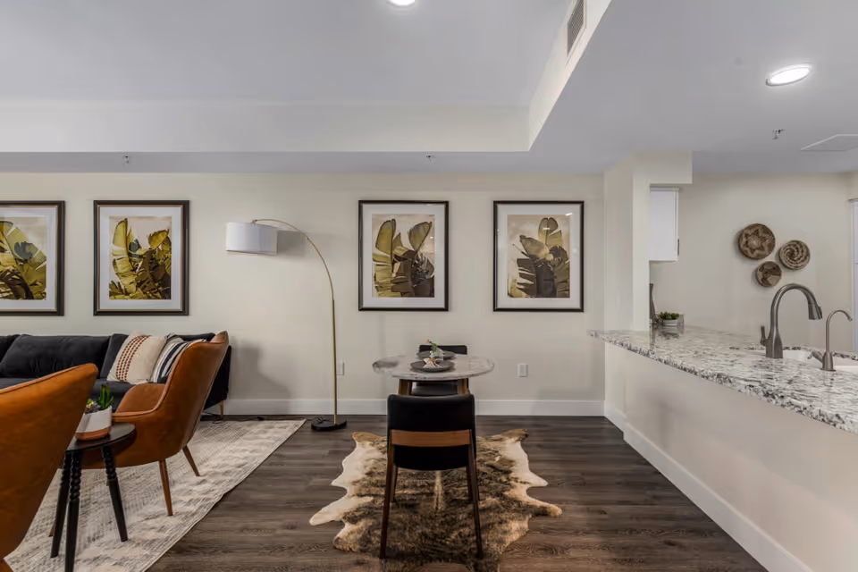 A modern living and dining area with a small round dining table and two chairs on a cowhide rug. The living room features a dark sofa with patterned pillows, two brown armchairs, a floor lamp, and three framed botanical prints on the wall. The kitchen counter with a granite countertop and a sink is visible on the right side, with decorative woven baskets on the wall.