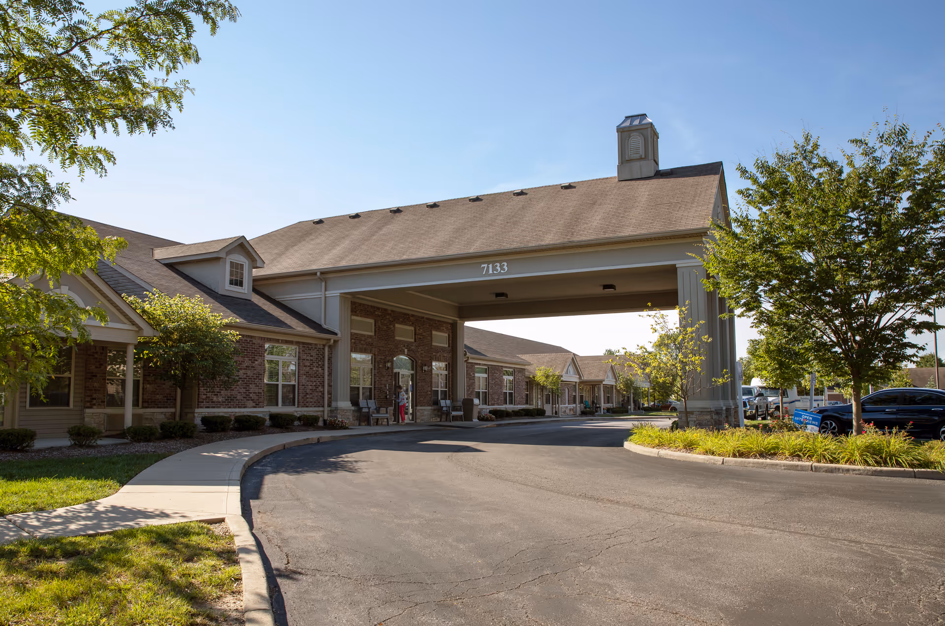 Entrance canopy and driveway of a single-story brick assisted living building with the number 7133 and landscaped grounds.