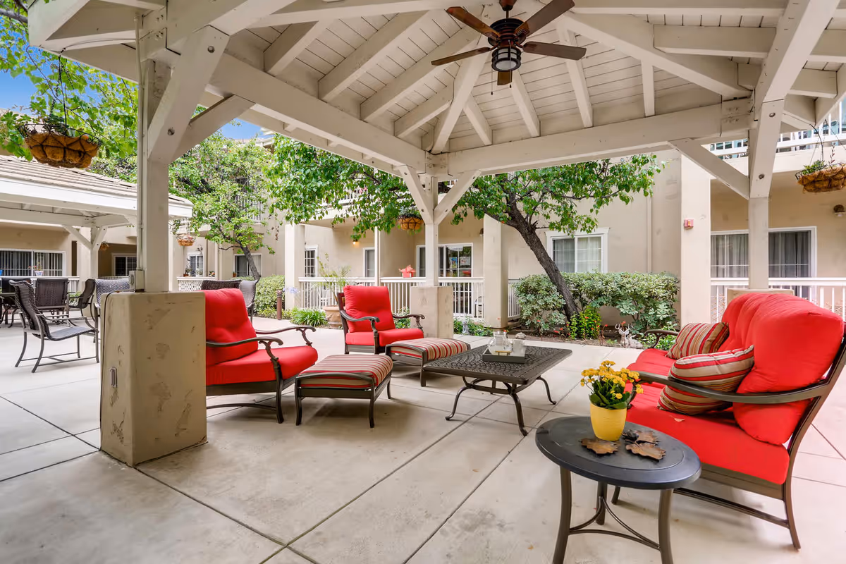 Outdoor covered patio area with red cushioned chairs and ottomans, a black metal coffee table, and a small round side table with a yellow flower pot. The patio is surrounded by greenery and beige building walls with windows.