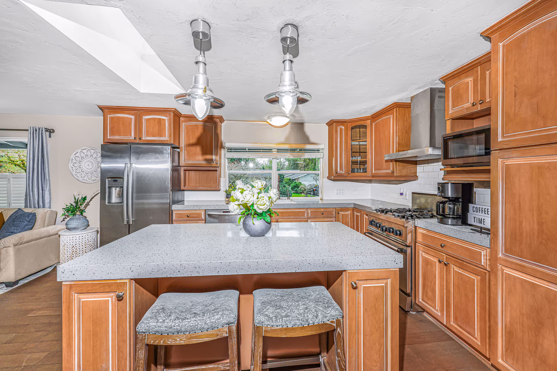 A bright kitchen with wooden cabinets, a large white speckled island countertop with two cushioned stools underneath, stainless steel refrigerator, stove, microwave, and coffee maker. A vase with white flowers is placed on the island. A window above the sink shows greenery outside, and part of a living room with a beige couch and blue pillows is visible to the left.