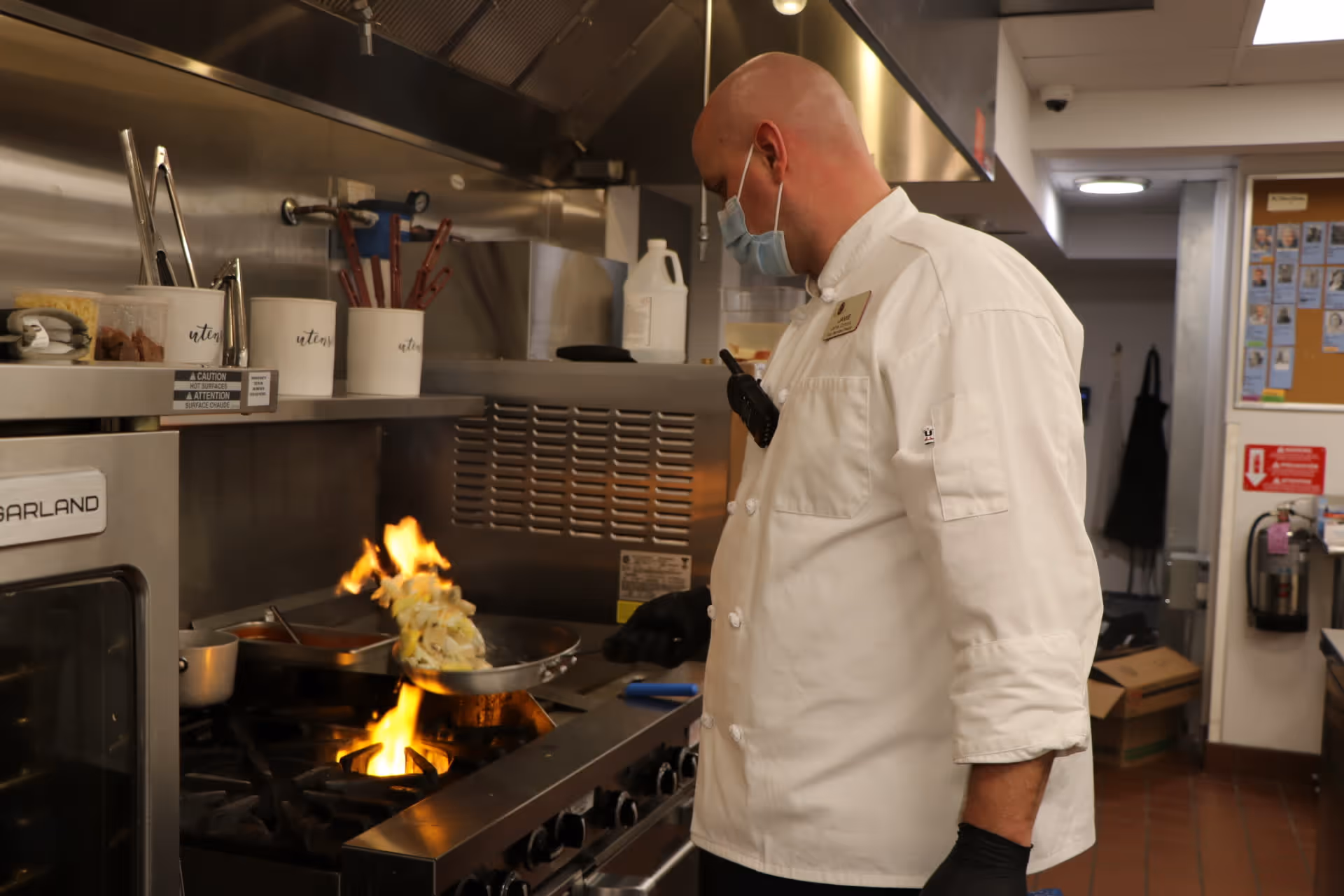 A chef wearing a white uniform, black gloves, and a face mask is cooking food in a stainless steel kitchen. The chef is holding a frying pan over a gas stove with visible flames, tossing the food in the pan. Various kitchen utensils and containers are visible on a shelf above the stove.