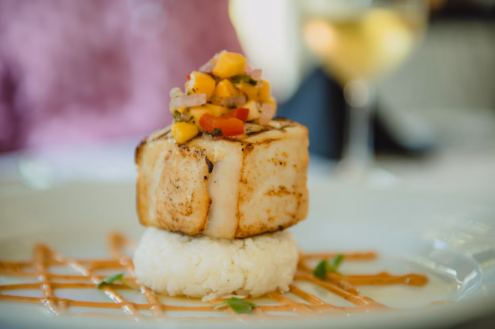 A seared square of fish served on a round rice cake topped with corn salsa and drizzled sauce on a white plate.