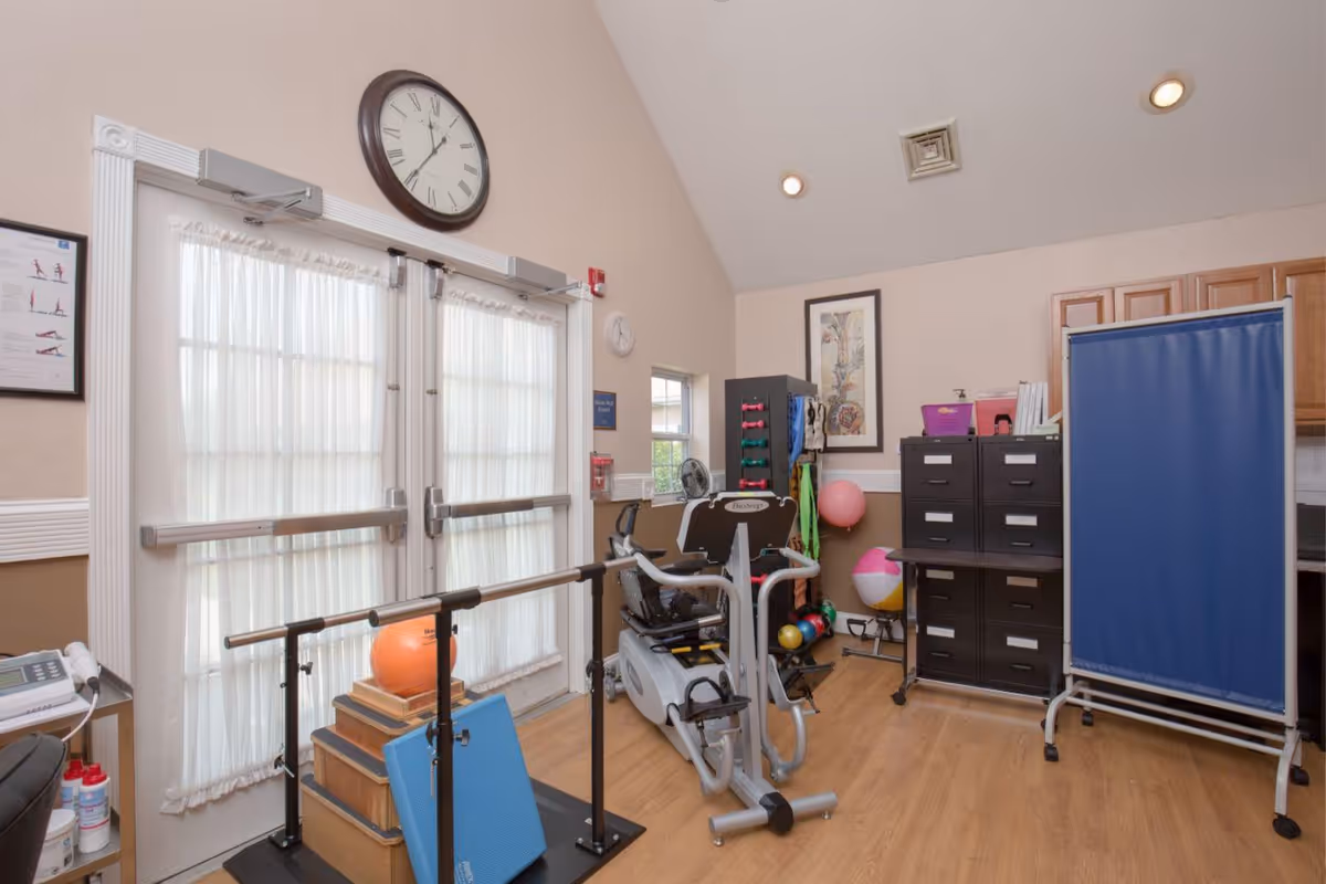 A small exercise room with various physical therapy equipment including a stationary bike, parallel bars, exercise balls, weights, and a blue privacy screen. The room has light-colored walls, wood flooring, a large clock above double doors with sheer curtains, and filing cabinets against the wall.