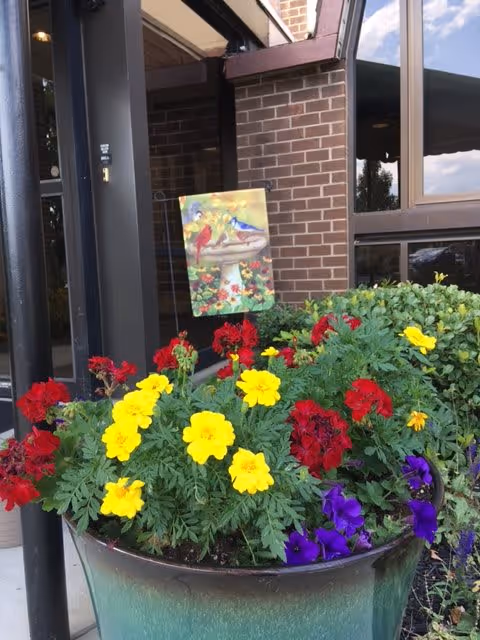 A large green planter filled with vibrant red, yellow, and purple flowers sits outside a brick building entrance. A decorative flag featuring colorful birds and flowers is placed in the planter. The building has large windows and a black metal door frame.