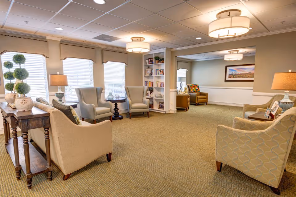 Well-lit communal sitting area with sofas and armchairs arranged around side tables and a bookshelf.