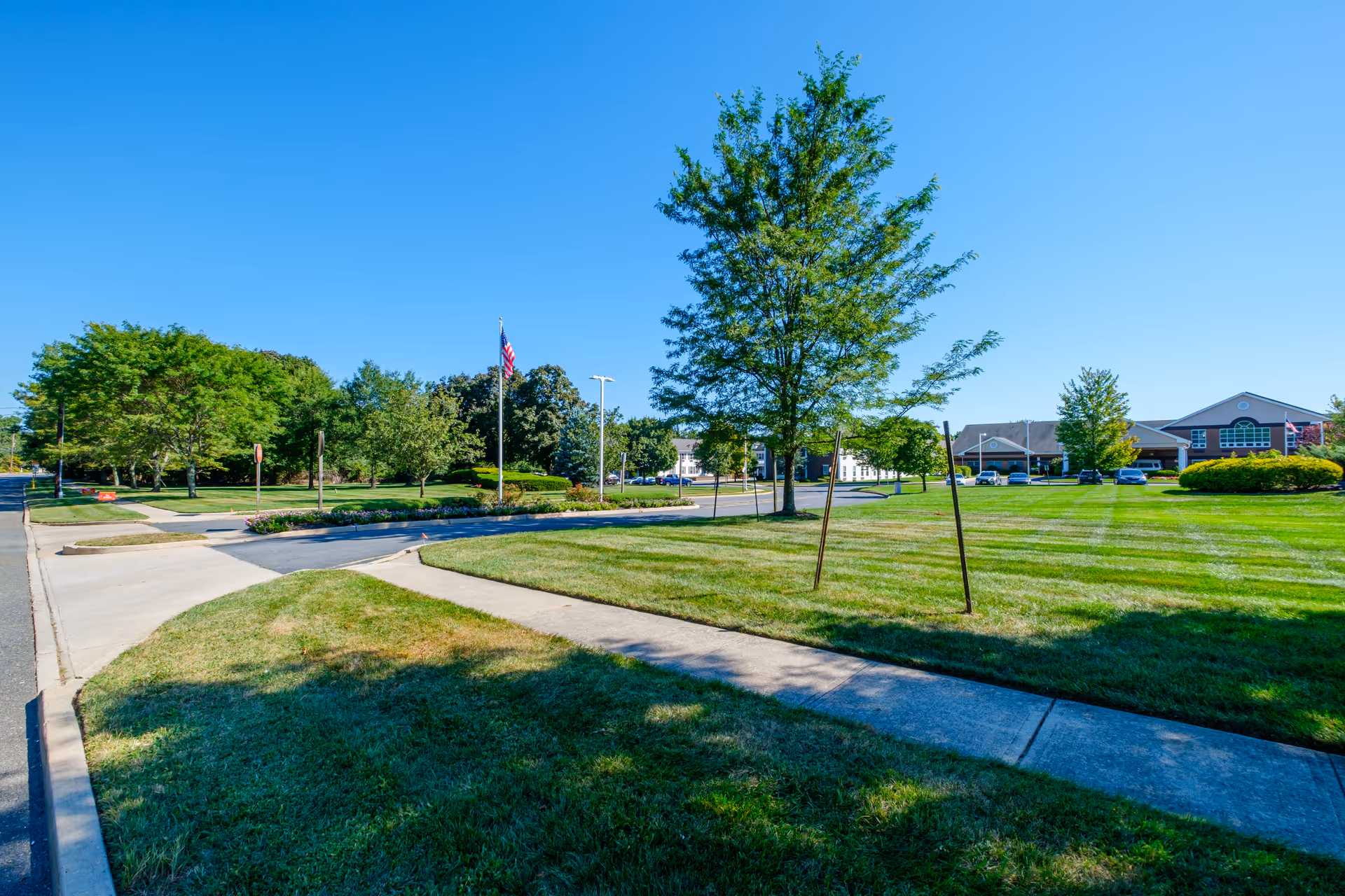 A sunny outdoor scene at Brandywine Toms River by Monarch showing a well-maintained lawn with a sidewalk, several trees, an American flag on a flagpole, and a building in the background under a clear blue sky.