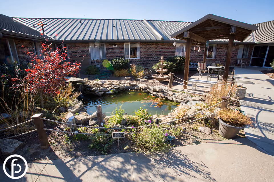 Outdoor courtyard area at LaVerna Manor Health & Rehabilitation featuring a small pond surrounded by rocks and plants, a wooden pergola with a table and chairs underneath, and a brick building with windows in the background.