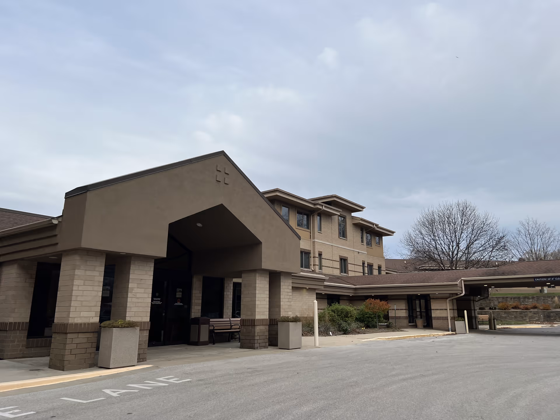 Exterior view of St Camillus Memory Care North Residence building entrance with a covered drop-off area, beige and brown brick facade, and a cloudy sky in the background.