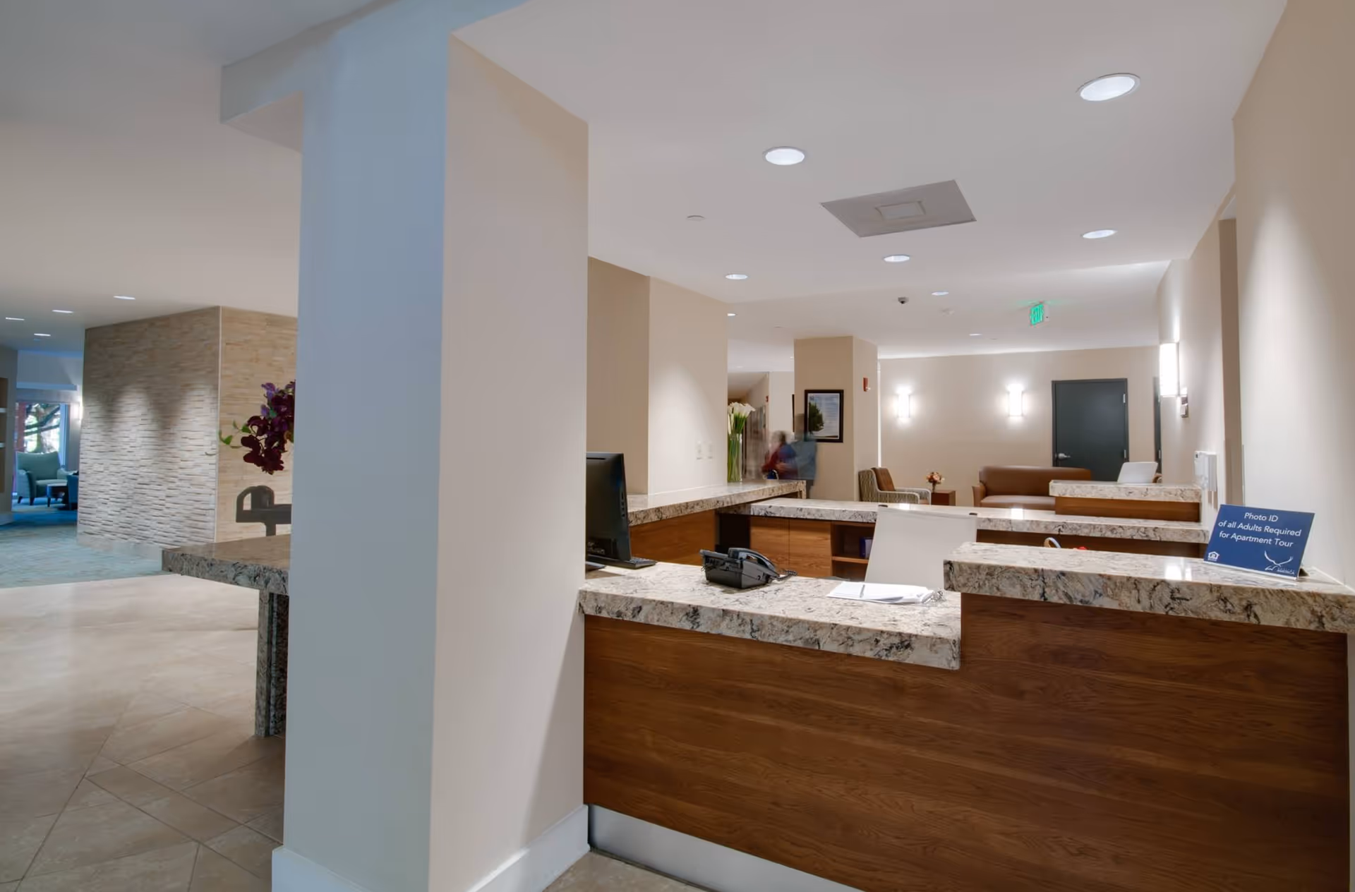 Lobby reception area with a marble-topped wooden front desk, seating area, and corridor under recessed lighting.
