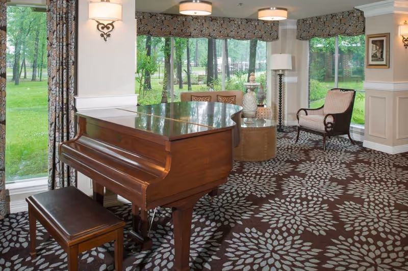 A cozy interior room with a polished wooden grand piano and matching bench on a patterned carpet. The room has large windows with floral curtains, allowing a view of green trees and grass outside. There is a comfortable armchair, a round ottoman, a floor lamp, and decorative wall sconces and artwork on the walls.