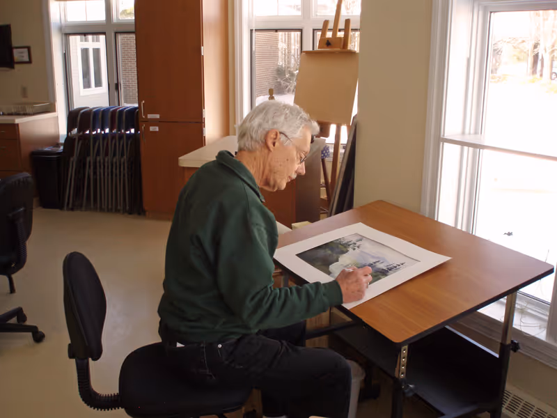 An elderly man with gray hair and glasses sits at a wooden table near large windows, working on a watercolor painting. The room is well-lit with natural light, and there are stacked chairs and an easel in the background.
