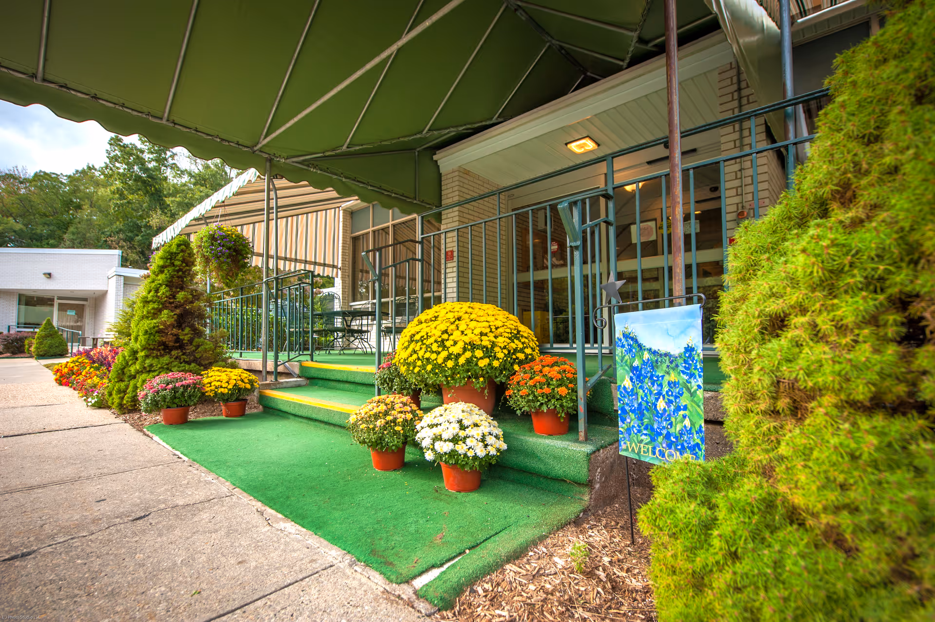 Entrance to a senior living facility with a green awning, steps and ramp lined with potted chrysanthemums and a welcome flag.