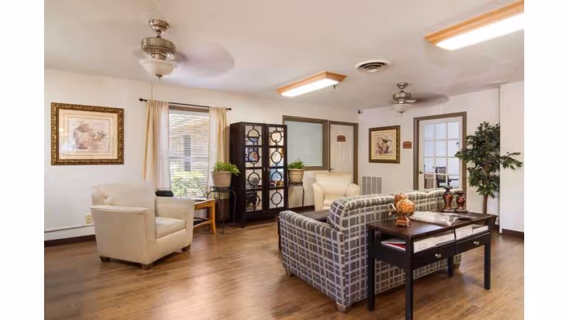 A cozy living room area in a nursing and rehabilitation center featuring a plaid sofa, two beige armchairs, a wooden side table with decorative items, a black display cabinet with glass doors, framed artwork on the walls, potted plants, and ceiling fans with lights. The room has wooden flooring and large windows with curtains allowing natural light.