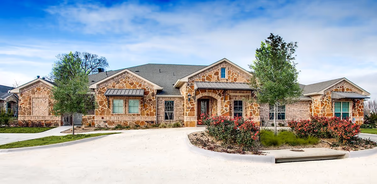 Front exterior view of Avalon Memory Care facility, a single-story building with stone and brick facade, multiple windows with awnings, surrounded by landscaped greenery including trees and bushes under a partly cloudy sky.