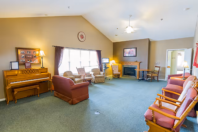 A spacious assisted living common area with a green carpet, beige walls, and a vaulted ceiling. The room features a wooden piano with a lamp and framed photos on top, several upholstered armchairs and wooden chairs arranged around the room, a fireplace with a framed picture above it, a ceiling fan with a light, and large windows with curtains allowing natural light to fill the space.