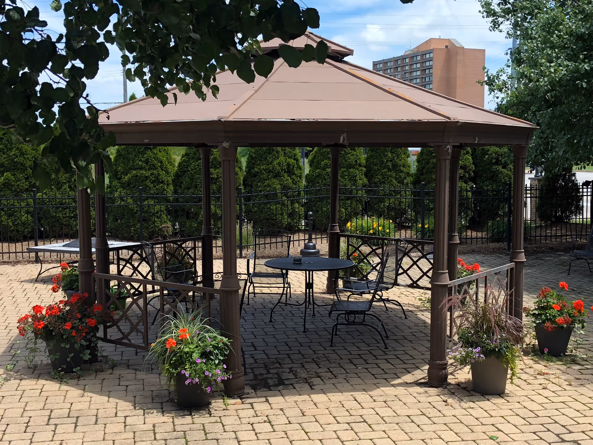 Outdoor gazebo with a brown roof and metal frame, surrounded by potted flowers with red and purple blooms. Inside the gazebo are black metal chairs and a round table on a paved patio. Green bushes and a black metal fence are visible in the background, along with a tall building in the distance.