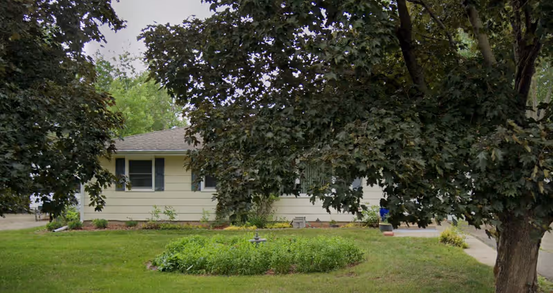 Single-story beige facility with a manicured lawn, shrubs, and large trees in front.