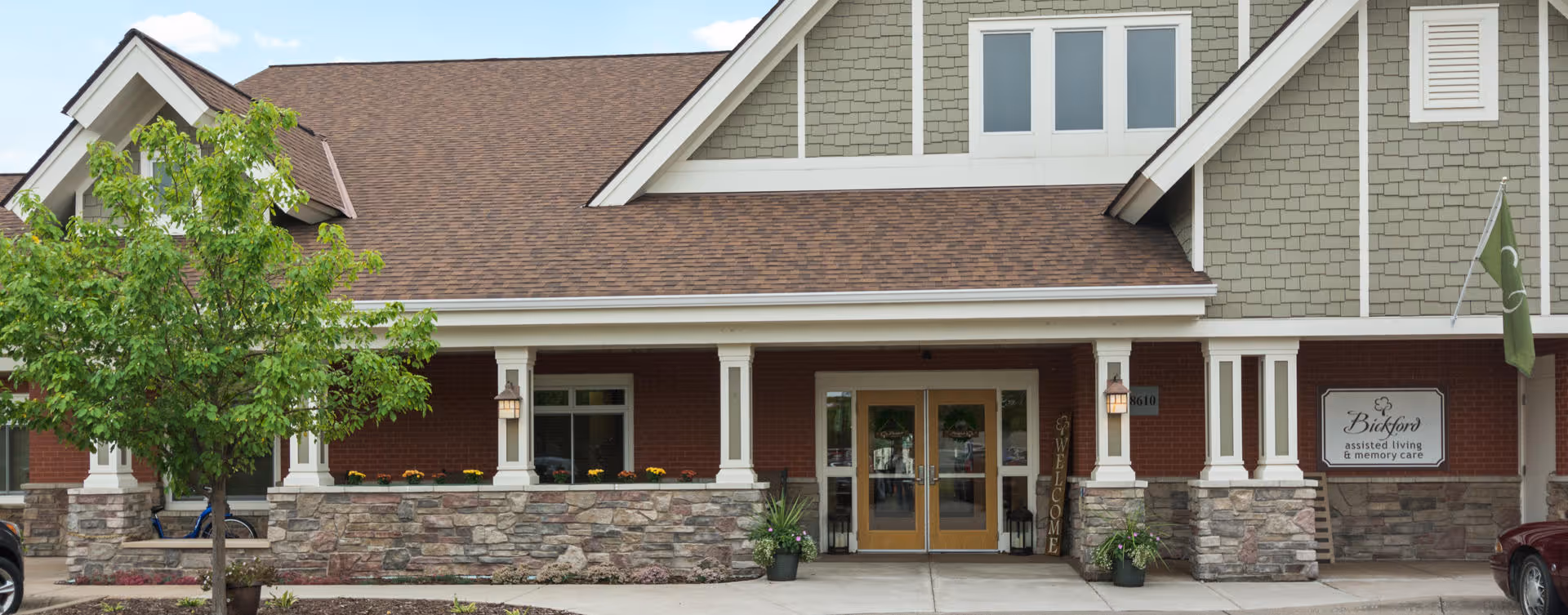 Exterior front view of a senior living facility building with a covered entrance, stone and brick facade, a tree on the left, and a sign that reads 'Bickford assisted living & memory care'.