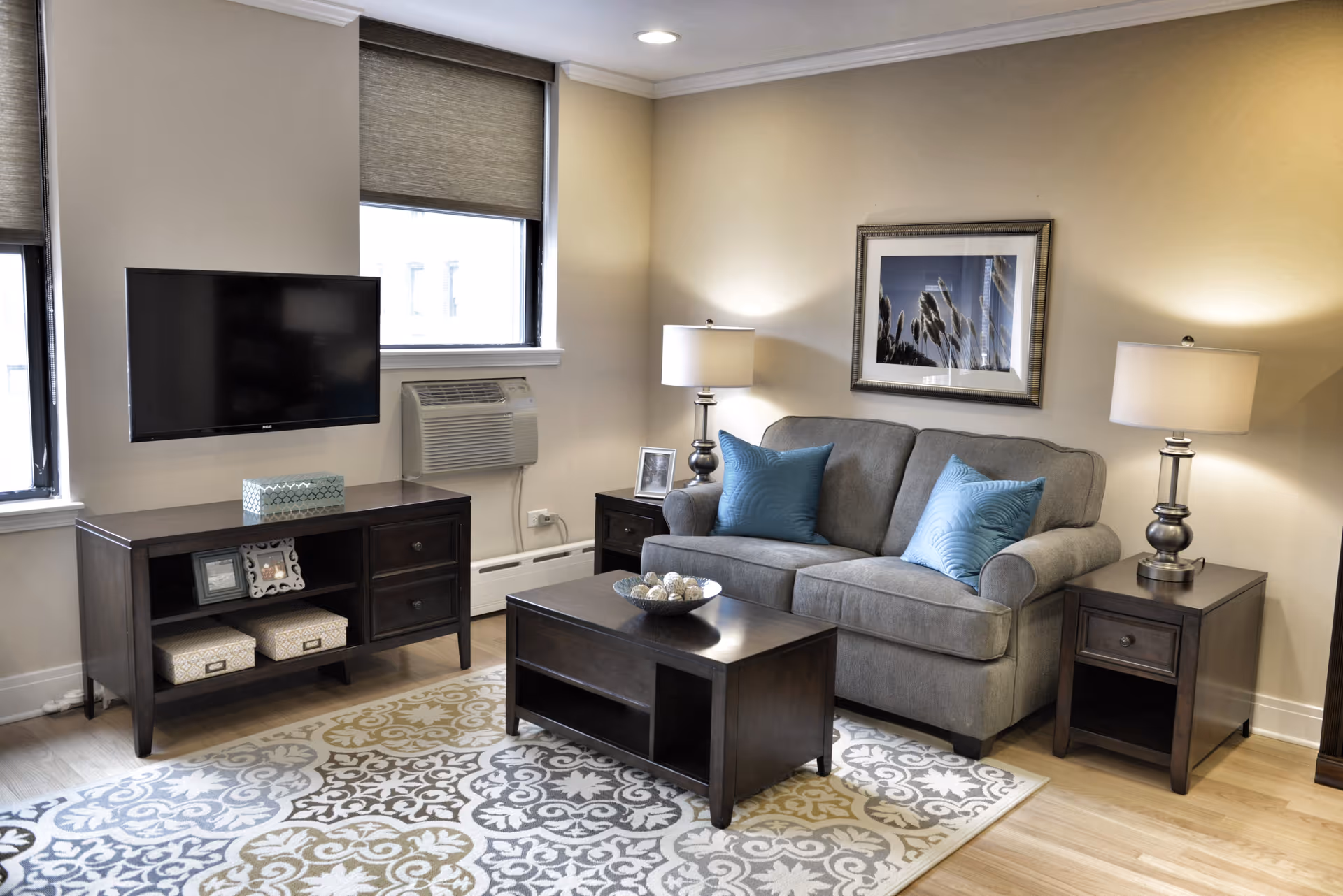 A cozy living room with a gray sofa adorned with two blue throw pillows, flanked by two matching dark wood side tables each with a lamp. A dark wood coffee table sits on a patterned area rug in front of the sofa. Across from the sofa is a dark wood TV stand with a flat-screen TV mounted on the wall above it. Two windows with beige blinds and a wall-mounted air conditioning unit are visible behind the TV stand. A framed picture hangs on the wall above the sofa.