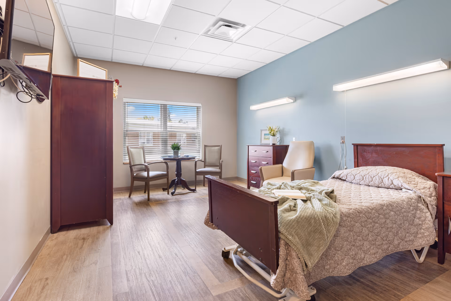 A well-lit senior living bedroom with a single bed covered in a beige quilt and a green blanket. The room features wooden furniture including a dresser, a nightstand, and a wardrobe. There is a beige armchair next to the dresser and a small round table with two chairs near a window with blinds. The walls are painted in light beige and blue tones, and two horizontal lights are mounted above the bed.