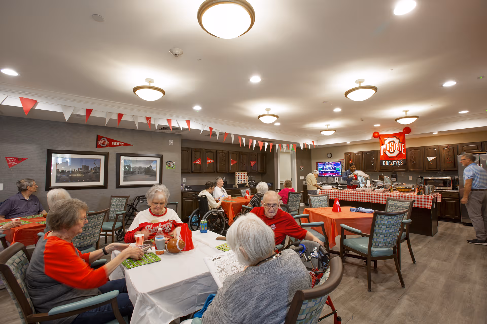 A group of elderly people sitting around tables in a decorated common dining area, playing games and socializing. The room has Ohio State Buckeyes themed decorations including banners, pennants, and a large sign on the wall. There is a kitchen area in the background with staff preparing food.