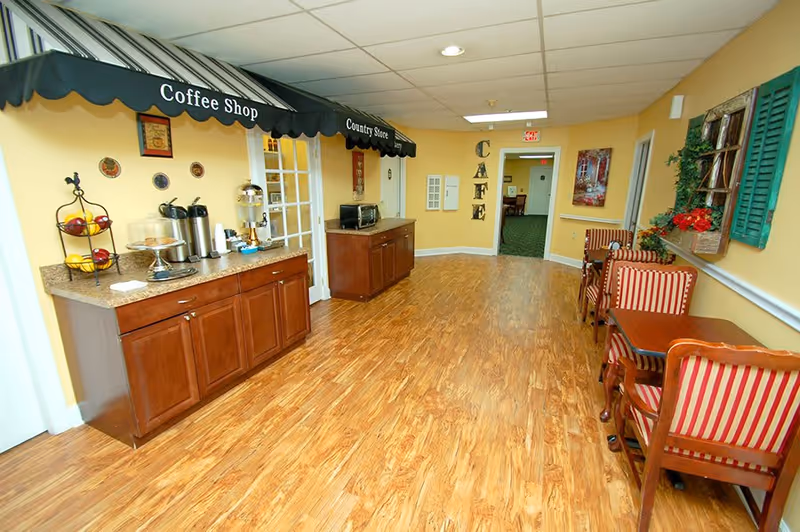 Bright indoor café area with a coffee station, wooden cabinets and small tables with striped chairs.