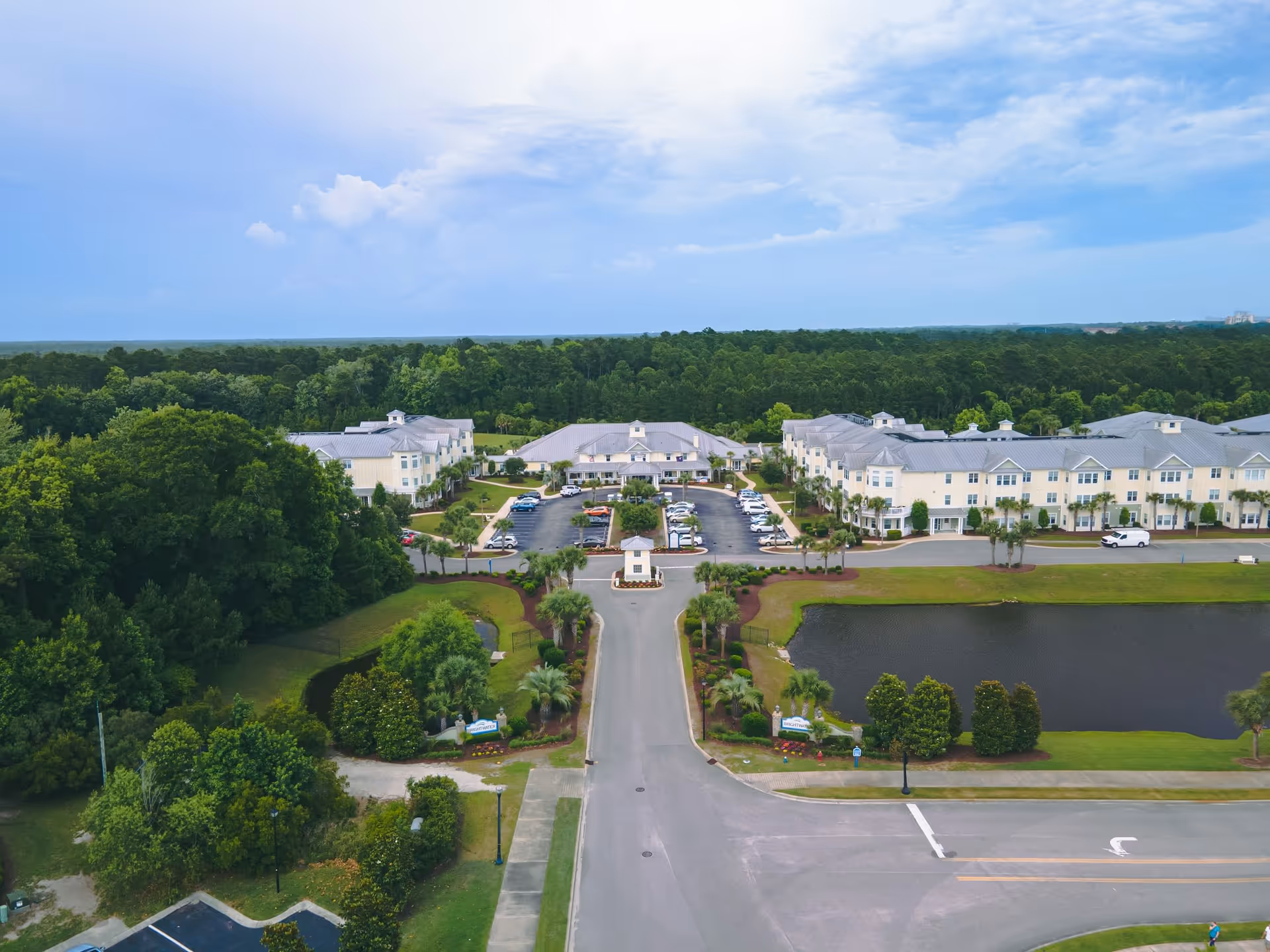 Aerial view of a senior living facility named Brightwater, showing multiple large buildings with light-colored roofs surrounded by greenery, a parking lot with cars, a pond, and a road leading to the entrance.