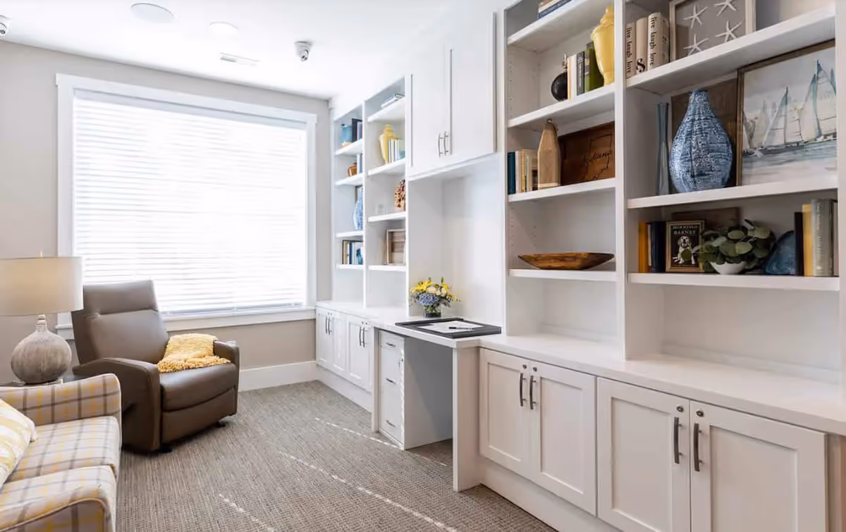 Bright sitting room with white built-in shelves and cabinets, a recliner, and a plaid sofa by a large window.