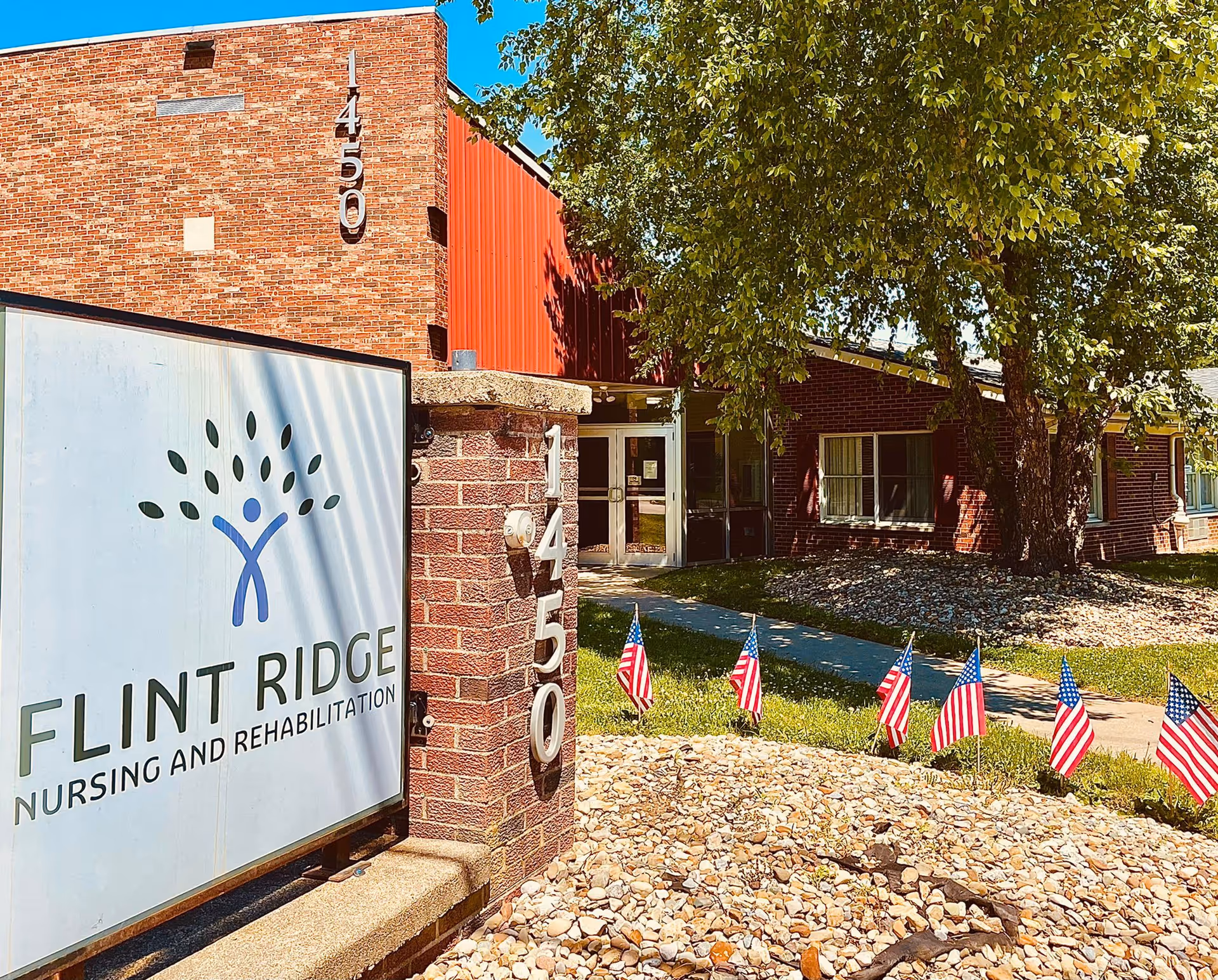 Exterior view of Flint Ridge Nursing and Rehabilitation facility showing a brick building with the address number 1450 displayed on a pillar and the building wall. There is a sign with the facility name and logo in front, a pathway leading to the entrance, several small American flags planted in the ground, and trees providing shade.